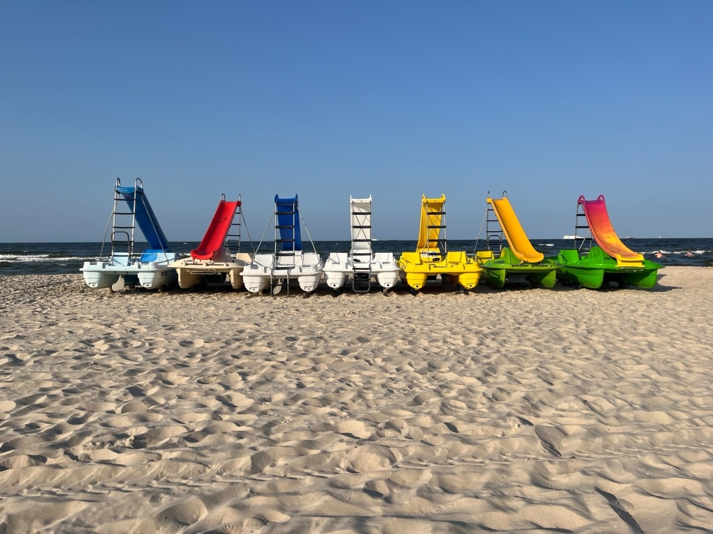 Verschiedenfarbige Treetbote  (Weiß, gelb, grün) mit Rutsche am Strand, etwas Meer im Hintergrund