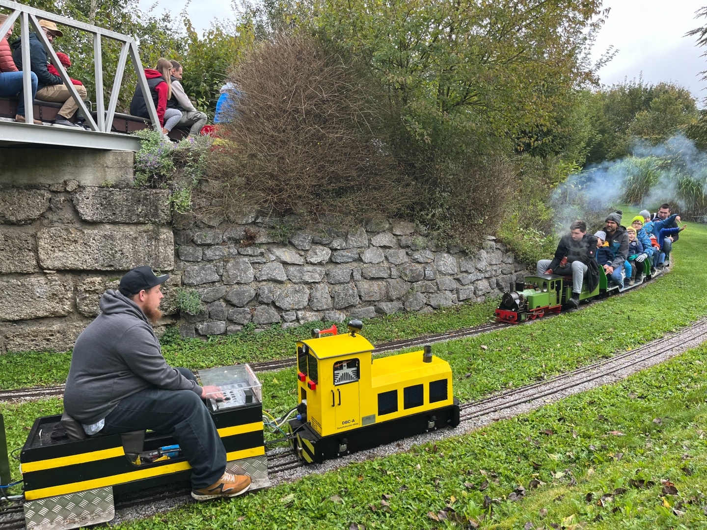 Eine gelbe Miniok und eine kleine grüne Dampflock fahren auf ihren jeweiligen Gleisen sich entgegen. Gleise auf grüner Wiese, man erkennt links oben auf einer Stein/Stahlbrücke einen dritten Minizug