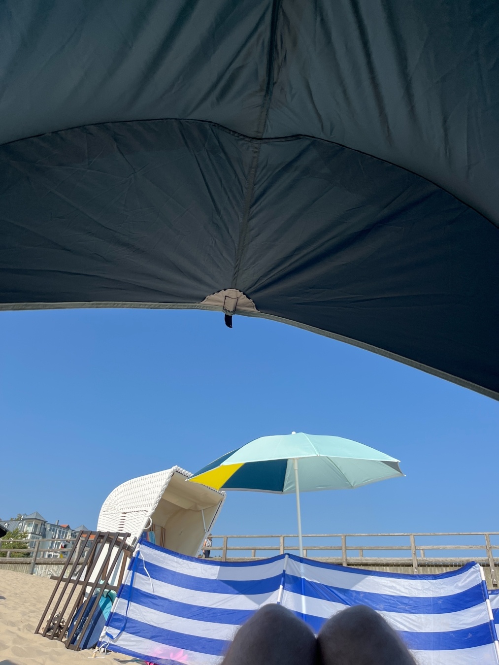 POV aus der Strandmuschel heraus. Man sieht deren grünes Dach und einen Strandkorb, vor dem ein weiß/blau gestreifter Windzaun gespannt ist.