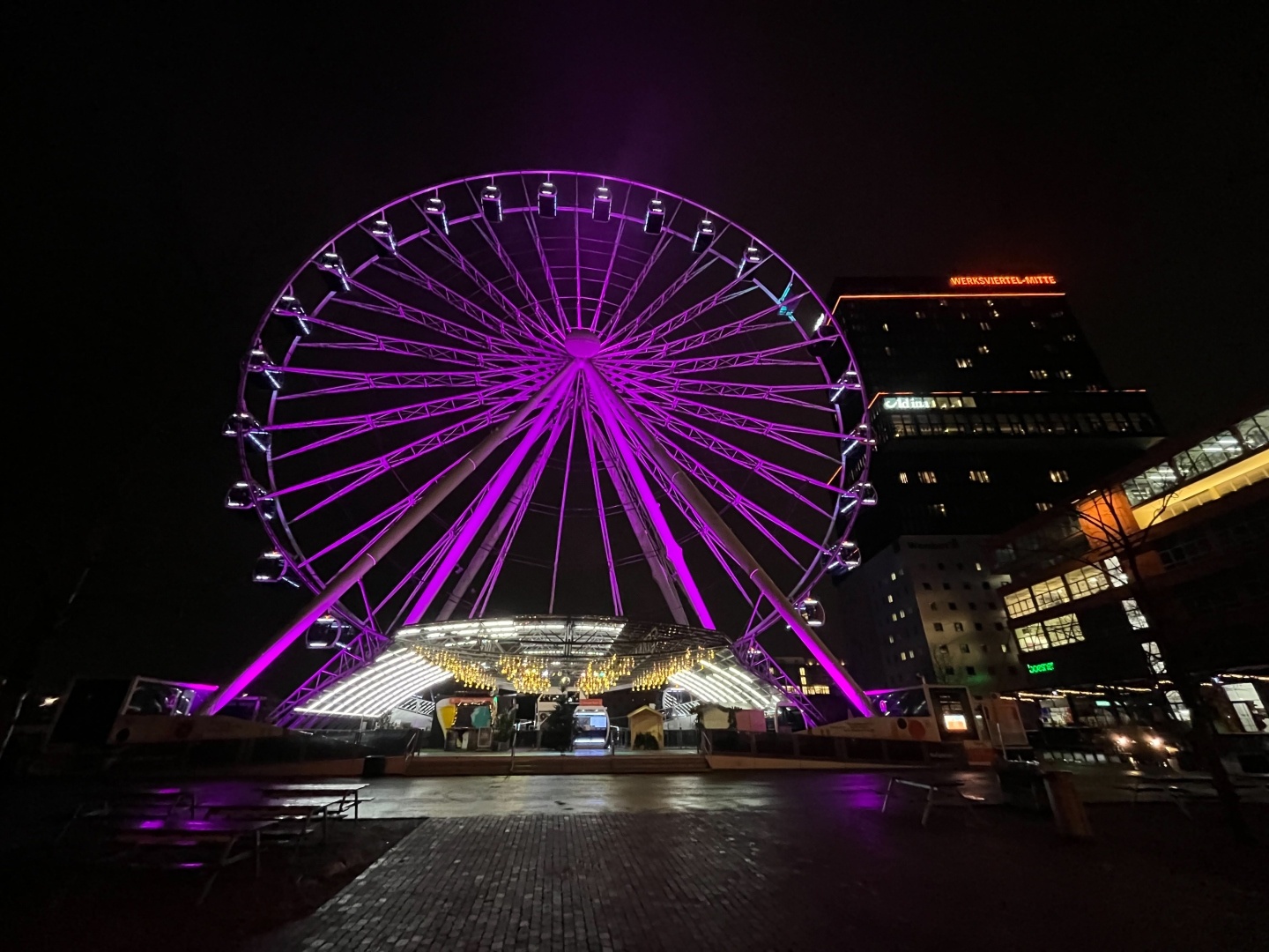 Lila/Violet angestrahltes Riesenrad, nebendran orange beleuchtetes Hochhaus mit oranger Schrift „Werksviertel Mitte“
Es ist dunkel/Nacht