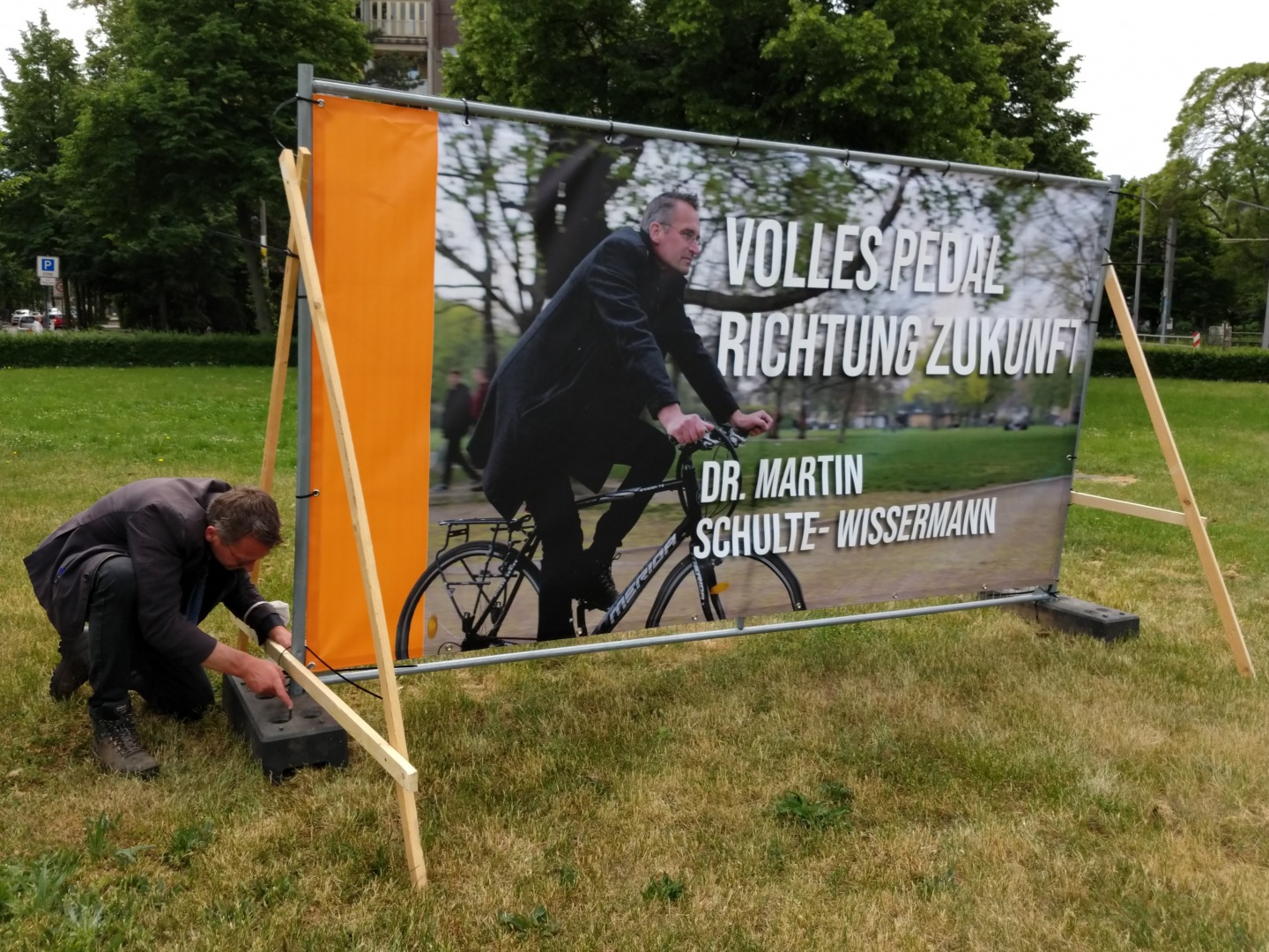 Martin kniet an einem Großplakat an einem Bauzaun und zurrt Kabelbinder fest. Auf dem Plakat ist er selbst auf einem Fahrrad abgebildet. Daneben die Worte: 