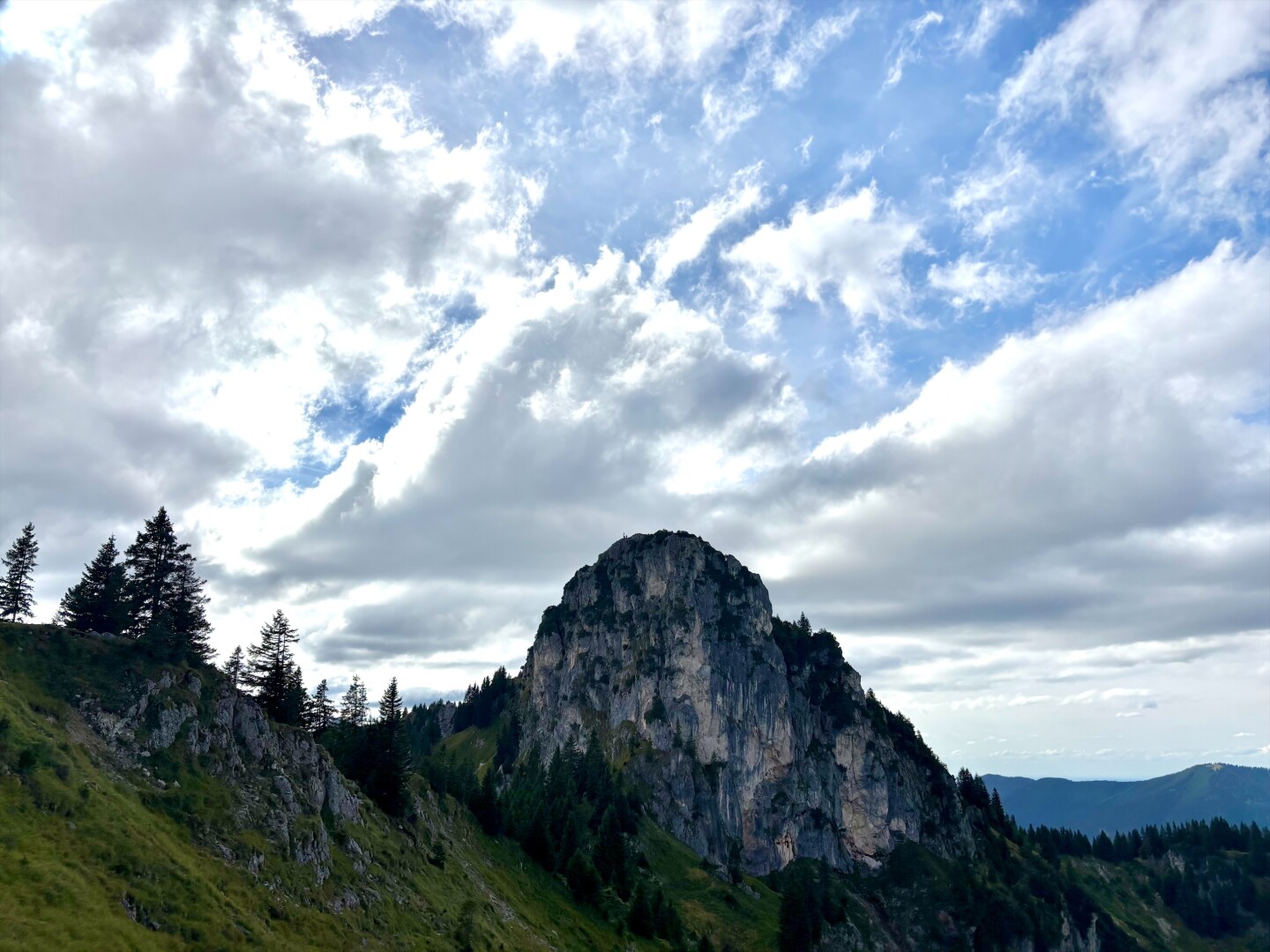 Ein felsiger Gipfel hinter einer steilen Wiese. Weiße Wolken am Himmel.