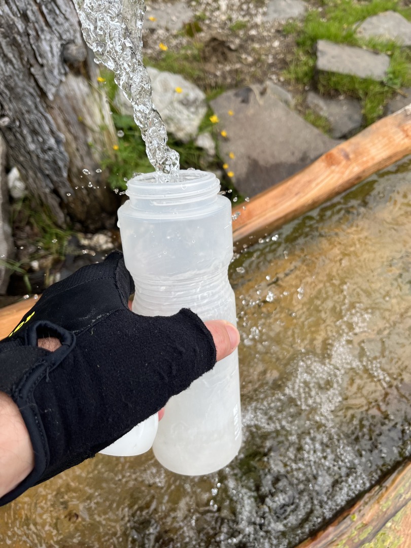 Eine Hand mit Handschuh hält eine Wasserflasche unter einen Brunnen
