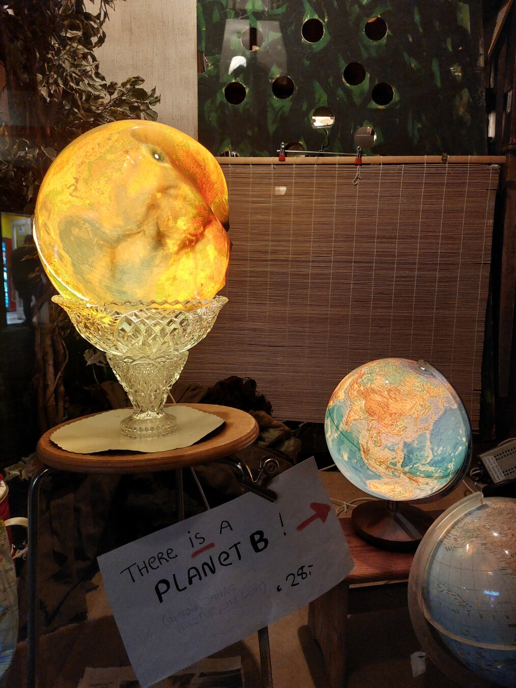 a window from a vintage store. 2 globes next to each other. the bigger one is melted and glows in orange light. Next to it is a smaller, intact globe. in front of them is a writing: there is a planet b! 28€