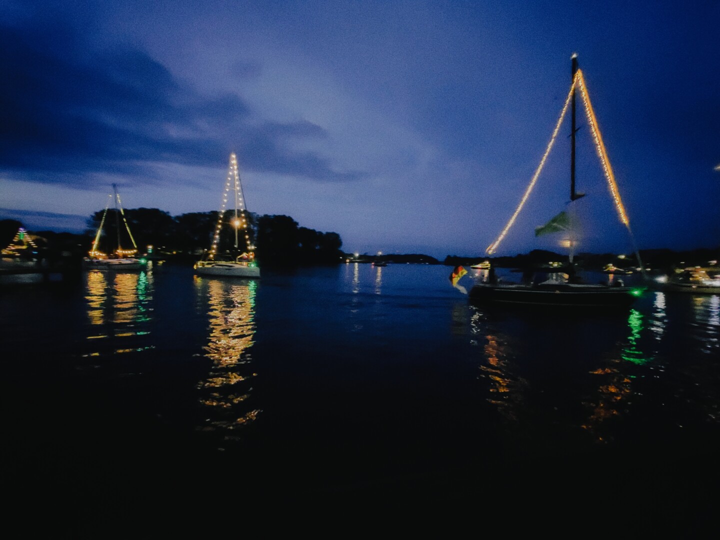 4 boats sailing in a row in the evening on dark water. All the boats have fairy lights on. The water is reflecting the lights. The sky is dark blue.