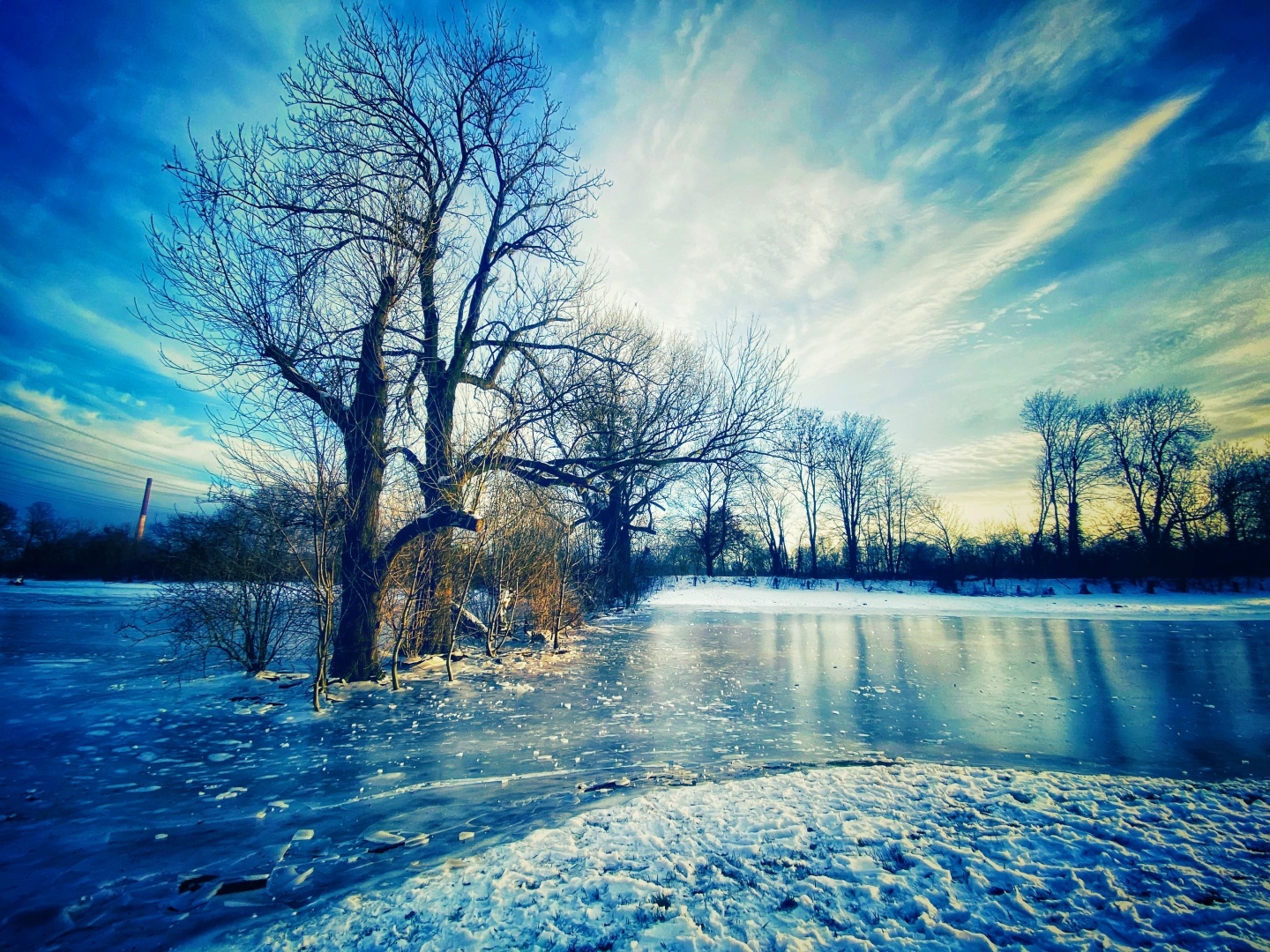 Ein winterliches Landschaftsfoto mit einem zugefrorenen See. Im Vordergrund stehen kahle, unbelaubte Bäume, während der Boden mit Schnee bedeckt ist. Der Himmel ist klar und zeigt sanfte Wolken in Blau- und Gelbtönen, die das Licht des Sonnenuntergangs reflektieren.