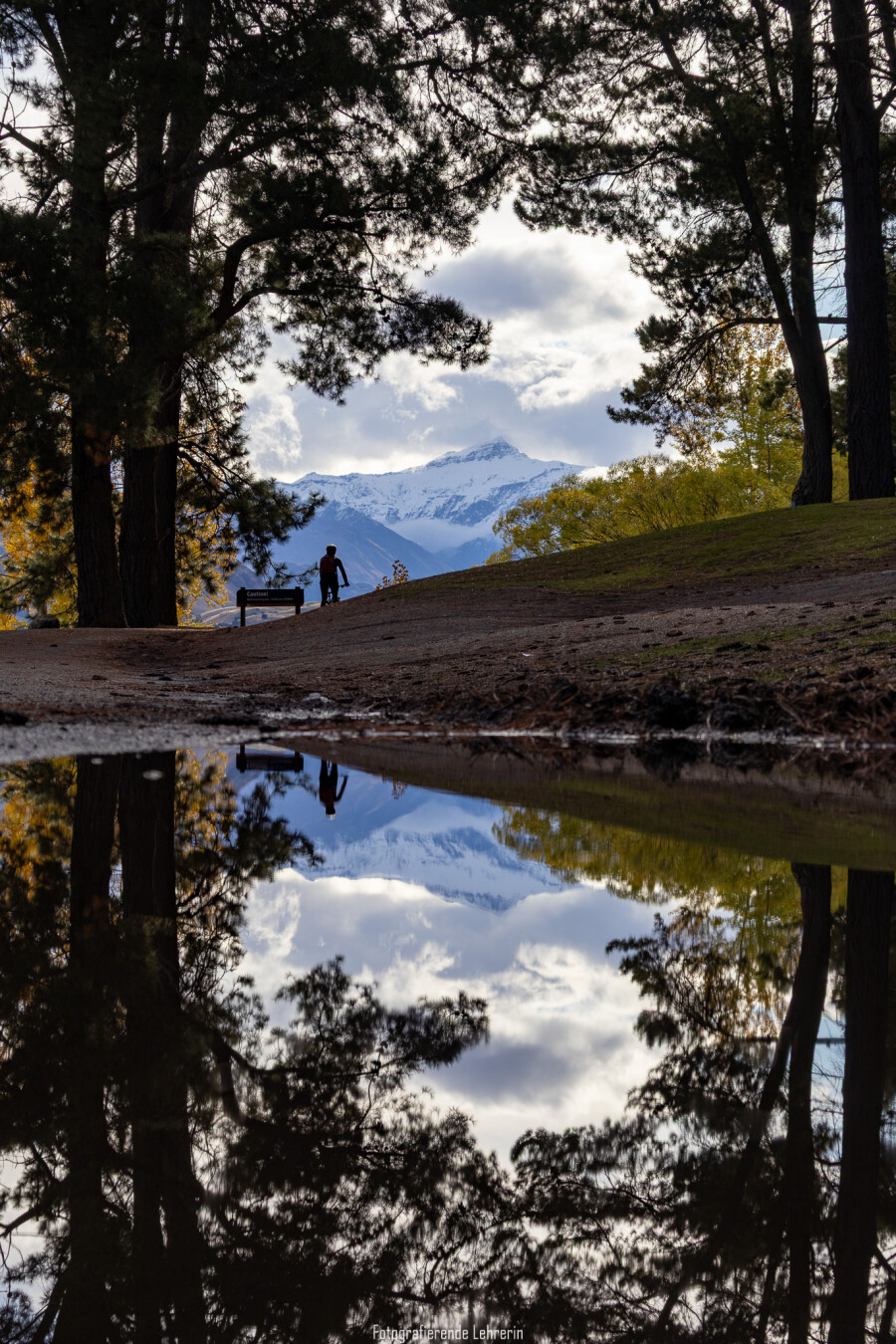Das Bild zeigt eine Parklandschaft mit hohen, dunklen Nadelbäumen, die links und rechts einen natürlichen Rahmen bilden. In der Bildmitte verläuft ein leicht ansteigender Weg. Darauf fährt ein Fahrradfahrer neben einem Schild. Im Hintergrund ist ein schneebedeckter Berggipfel zu sehen, teilweise von Wolken umgeben. Das Licht kommt von vorne, wodurch Bäume und Person dunkel wirken. Im Vordergrund befindet sich eine größere Wasserpfütze, in der sich die Bäume, die Wolken, der Berg und die Person nahezu spiegelbildlich reflektieren.