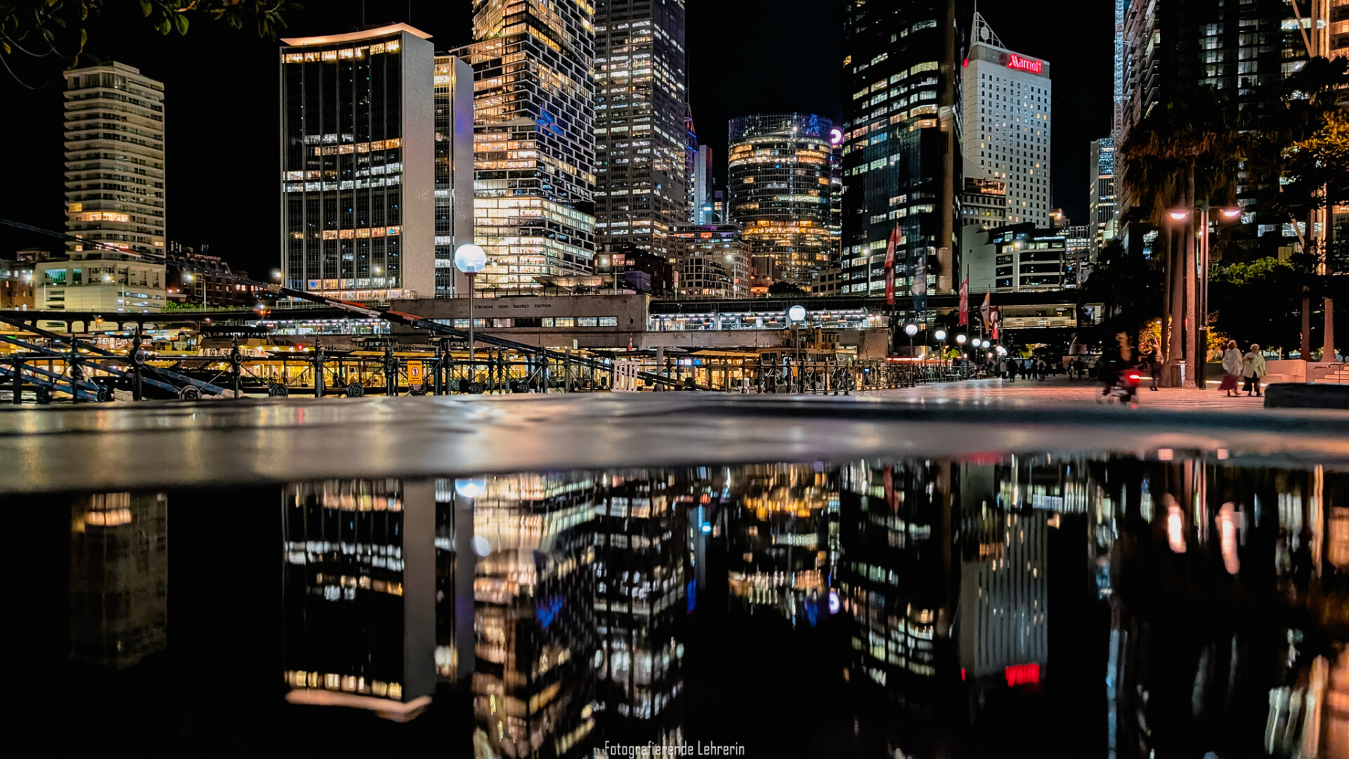 Das Bild zeigt eine moderne Großstadt bei Nacht mit zahlreichen beleuchteten Hochhäusern an einer Hafenpromenade. Die Lichter der Gebäude spiegeln sich in einer großen Wasserpfütze im Vordergrund, wodurch der Eindruck eines zweiten, spiegelverkehrten Stadtpanoramas entsteht. Entlang der Uferpromenade sind Lampen, Geländer, einige Passanten und mehrere Palmen zu sehen. (Beschreibung durch KI: Microsoft Copilot)