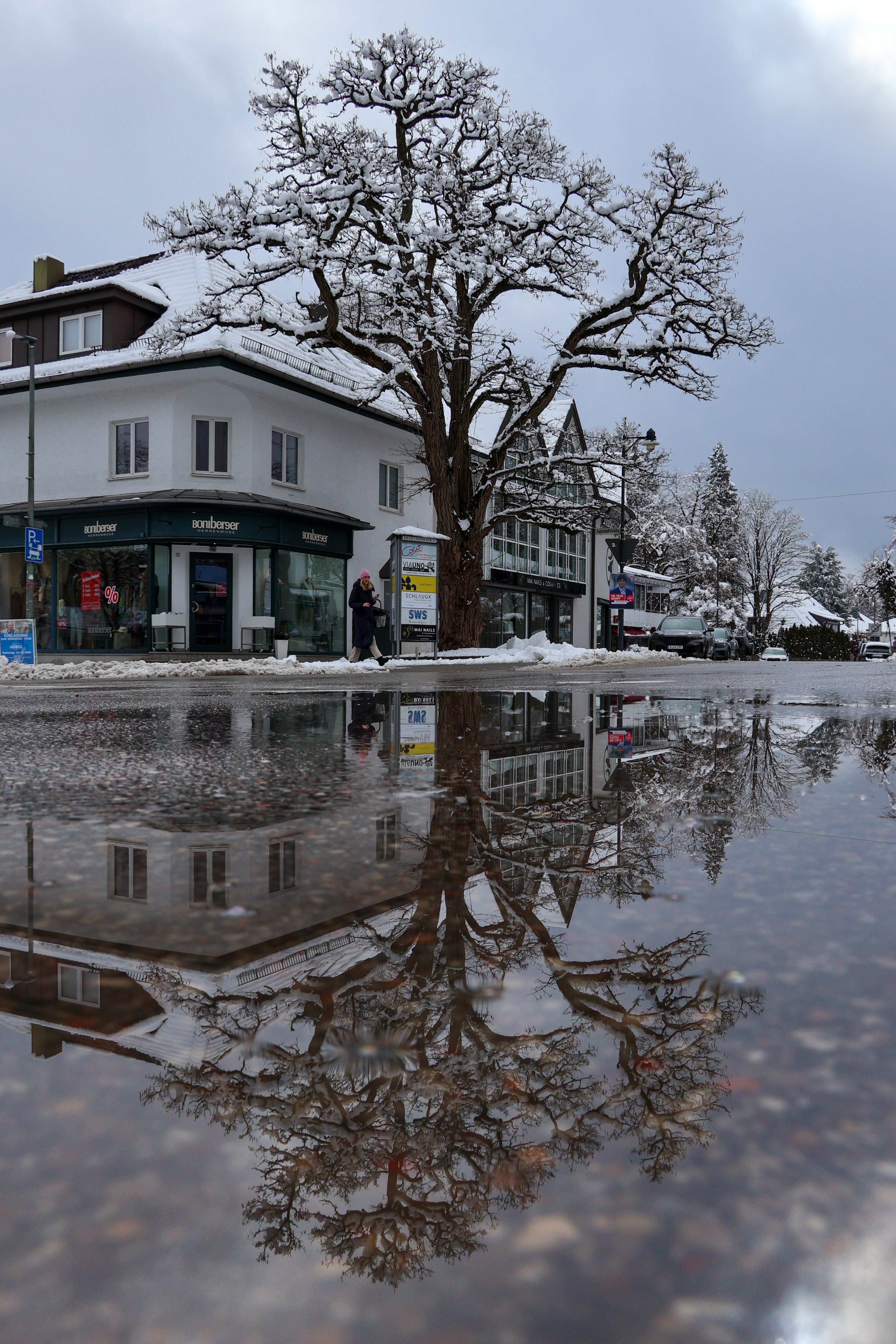 In einer verschneiten Stadtstraße steht ein großer, kahler Baum im Vordergrund, dessen Äste von Schnee bedeckt sind.

Dahinter befindet sich ein mehrstöckiges

Eckhaus mit einem Laden im Erdgeschoss,

vor dem eine Frau mit rosa Mütze auf dem

schneebedeckten Gehweg vorbeigeht.

Auf der Straße liegt eine große Pfütze, in der sich sowohl der Baum als auch die Gebäude klar spiegeln. Die Szene wirkt ruhig und winterlich, der Himmel ist grau und bedeckt.