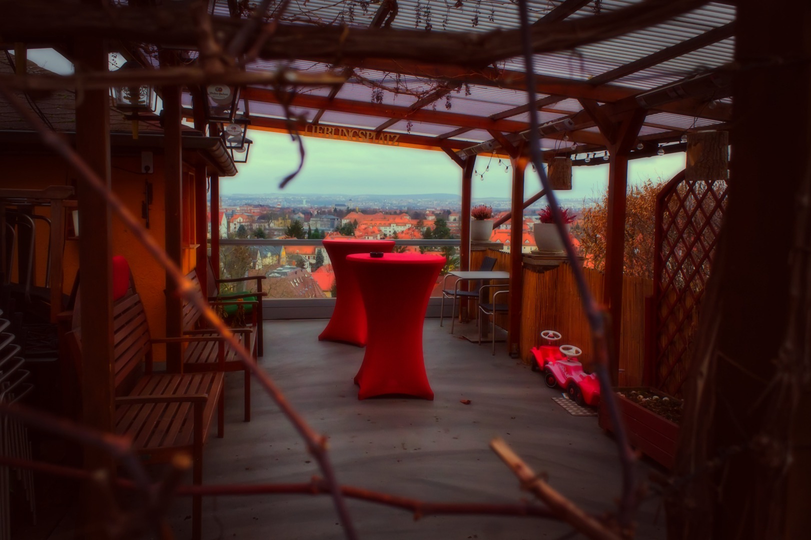 A shot of a terrasse of a restaurant in Dresden. The terrasse is nearly empty, some benches are on the side but there are two tall desks enclad with bright red fabric. Behind, the view opens over the city of Dresden. On one of the bars of the terrasse root, the word "Lieblingsplatz" is shown (which translates to "favourite place").