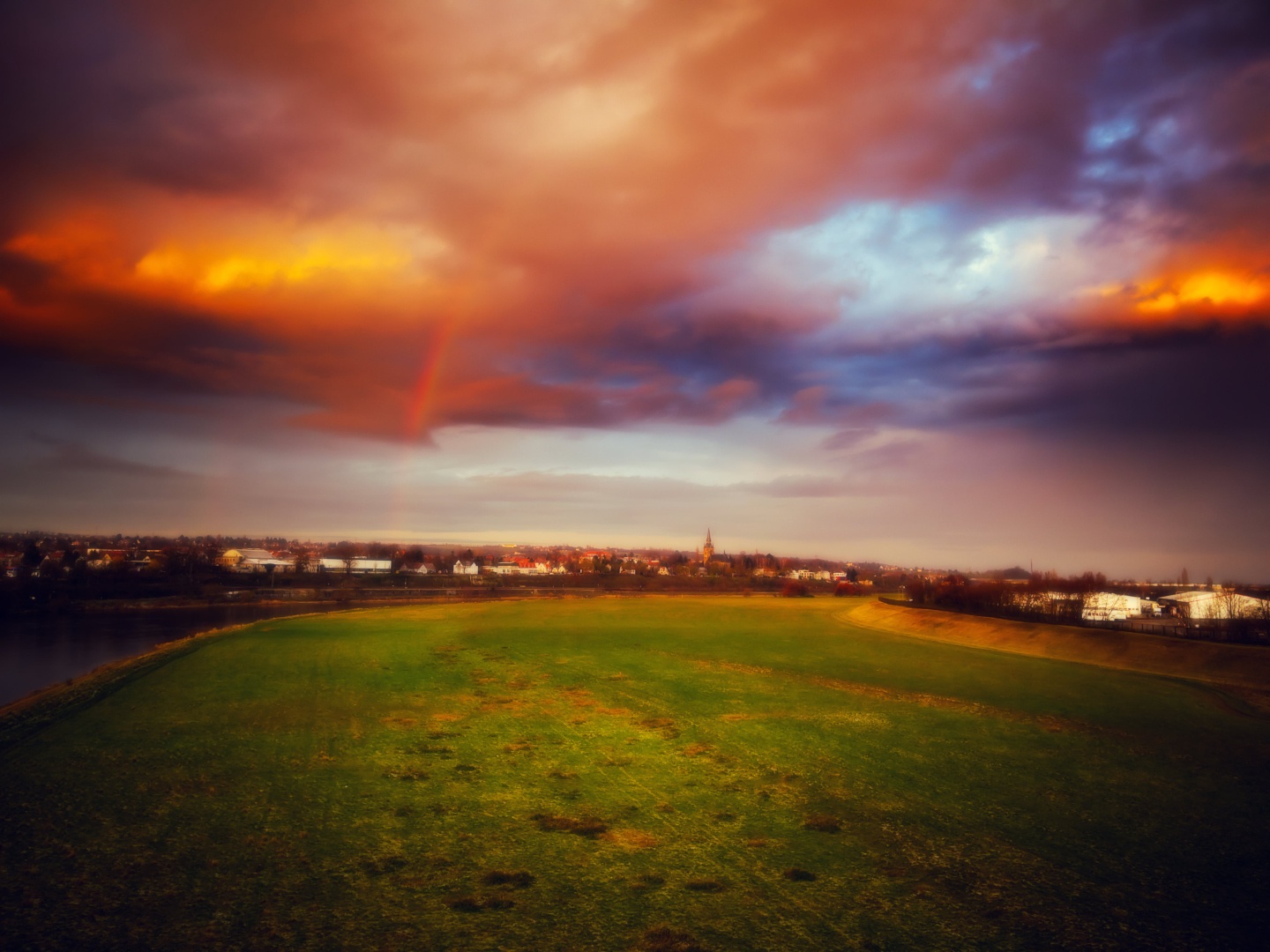 A view onto the lawns at the Elbe river. It is morning, the sun (behind the photographer) illuminates the clowds in beautiful orange tones. A tiny piece of a rainbow is also visible, indicating it is going to start raining soon.