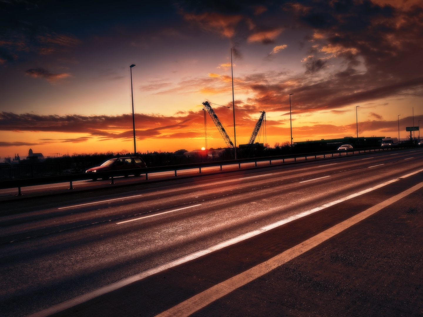 The picture shows a road during sunrise. The sky and clouds are cast in a nice, orange color. Some cars are currently passing by. In the background, a crane can be seen, next to which the sun just crossed the horizon.