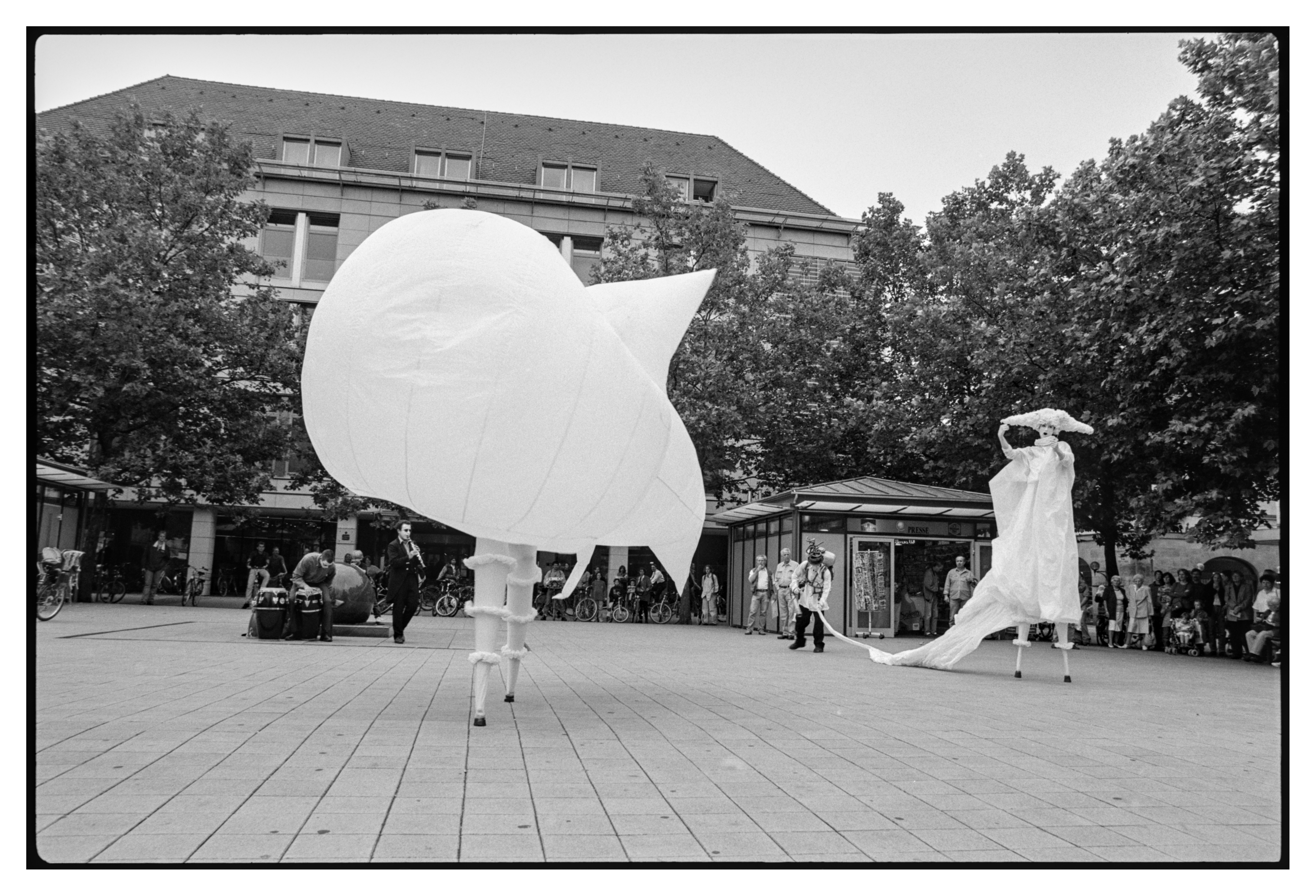 Black and white photograph in landscape orientation.
Another actor has entered the scene and is inflating the costumes of two of the original actors. The first one is now wear a balloon like costume with 2-3 meters diameter. The second one is being filled up.
