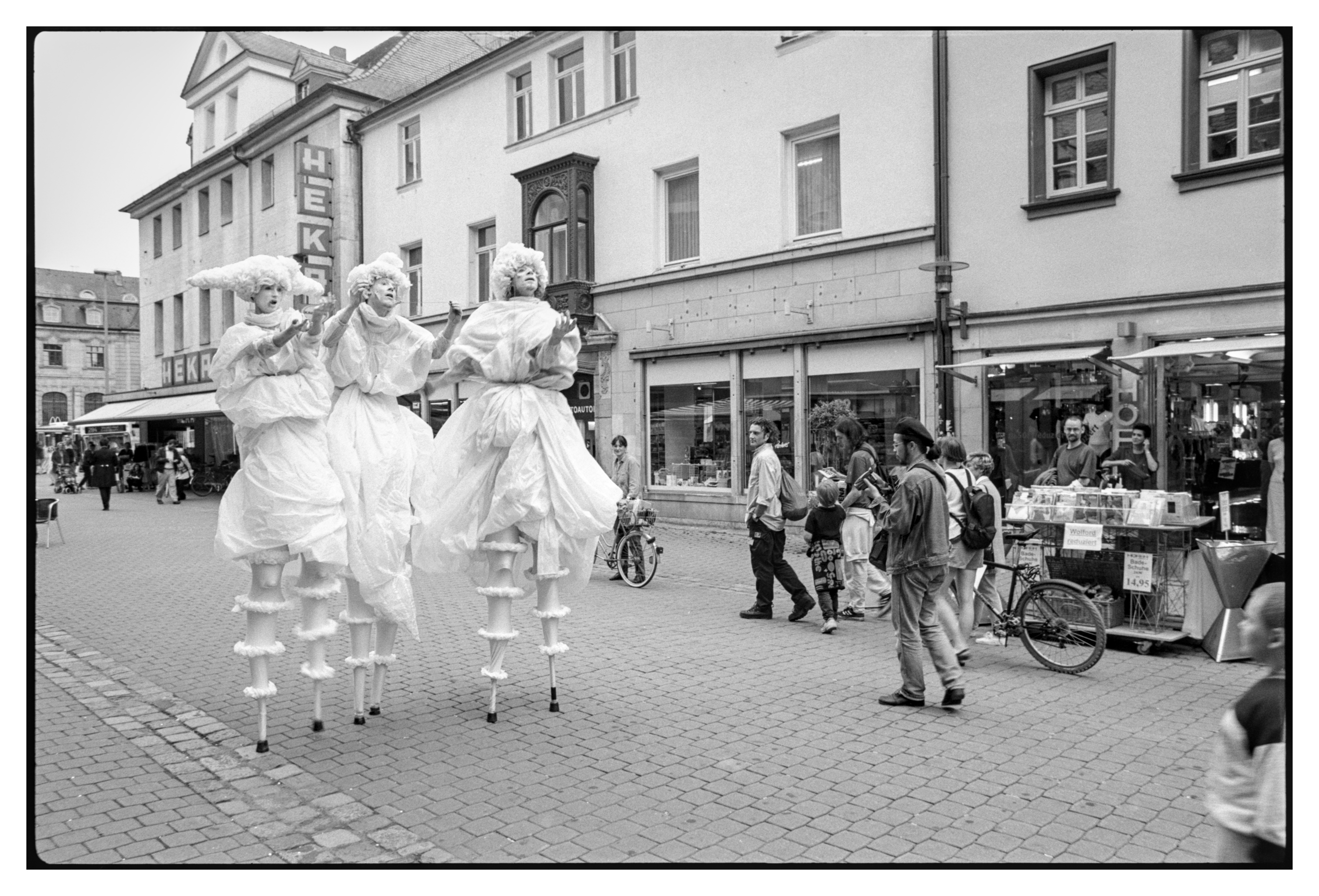 Black and white photograph in landscape orientation.
Three actors on stilts with white costumes are standing in a street with some people looking interested while others are minding their own business.
