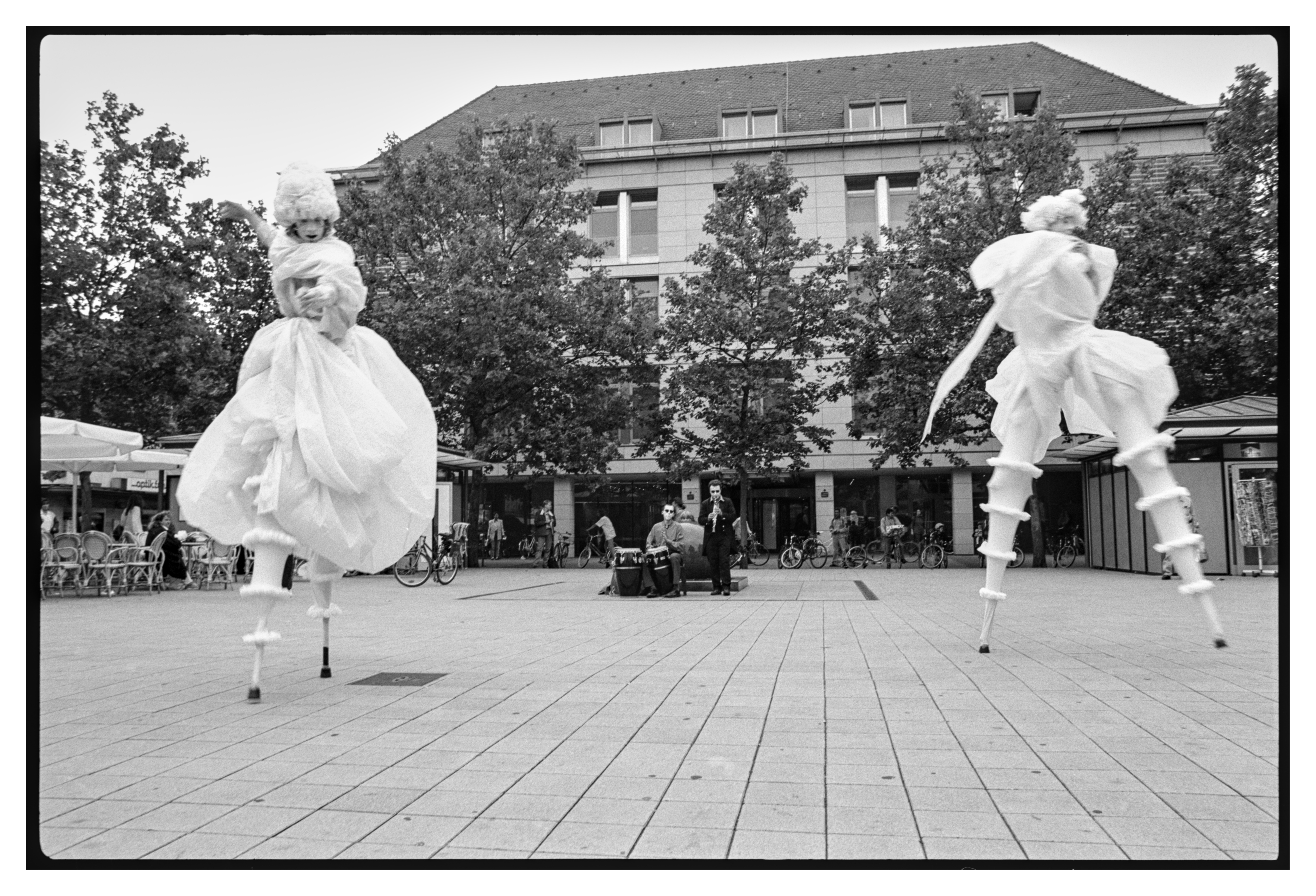 Black and white photograph in landscape orientation.
Two actors are using their stilts to make really large steps on a city square. Musicians but also normal street live in the background.
