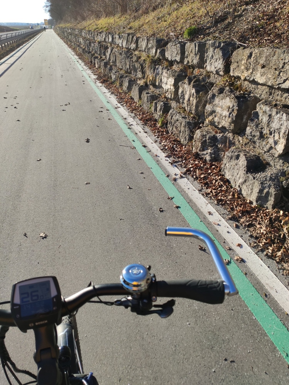 Man sieht im Vordergrund einen Fahrradlenker und dahinter einen schnurgeraden Radschnellweg mit einer Mauer rechts daneben. Es ist deutlich hell-sonnig.