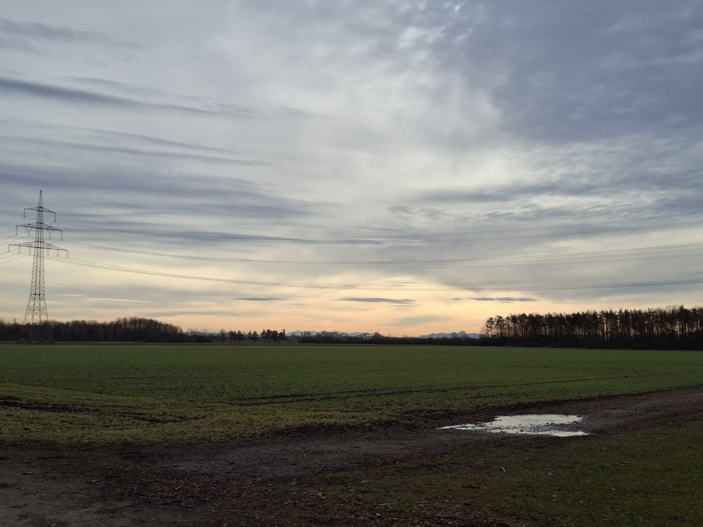 A rural landscape at dawn. You can see a puddle a power pylon, and woods and montains in the far out.