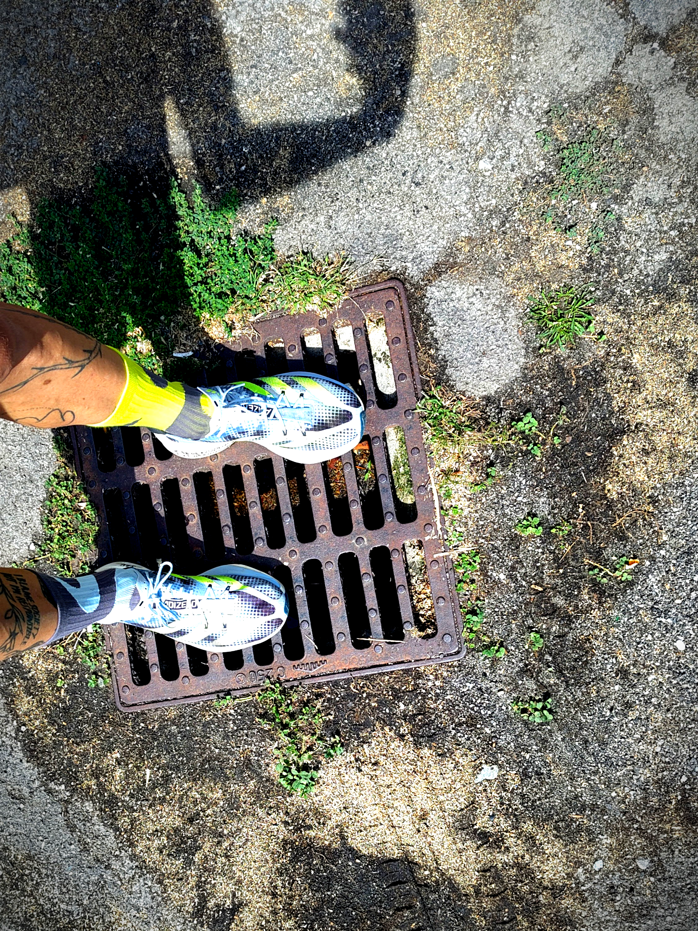 a photo from the floor which is broken up and has plenty of little plants growing, a sewer metal drain and feet and legs as well as a hard shadow of photographer legs and arms