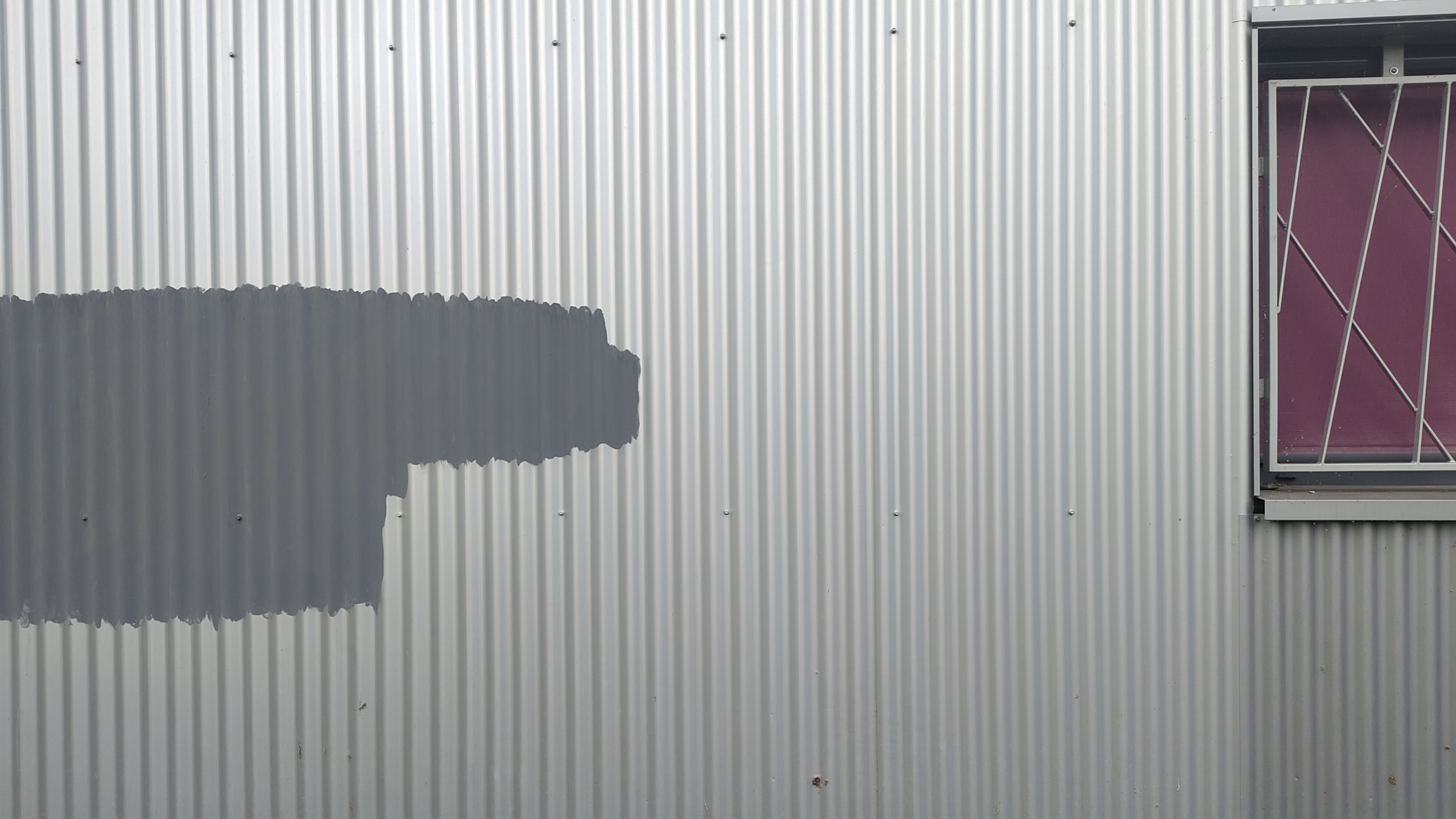 a grey metal wall with a darker grey part where the paint seems to have been removed. on the right corner a window can be partially seen with dark purple shades