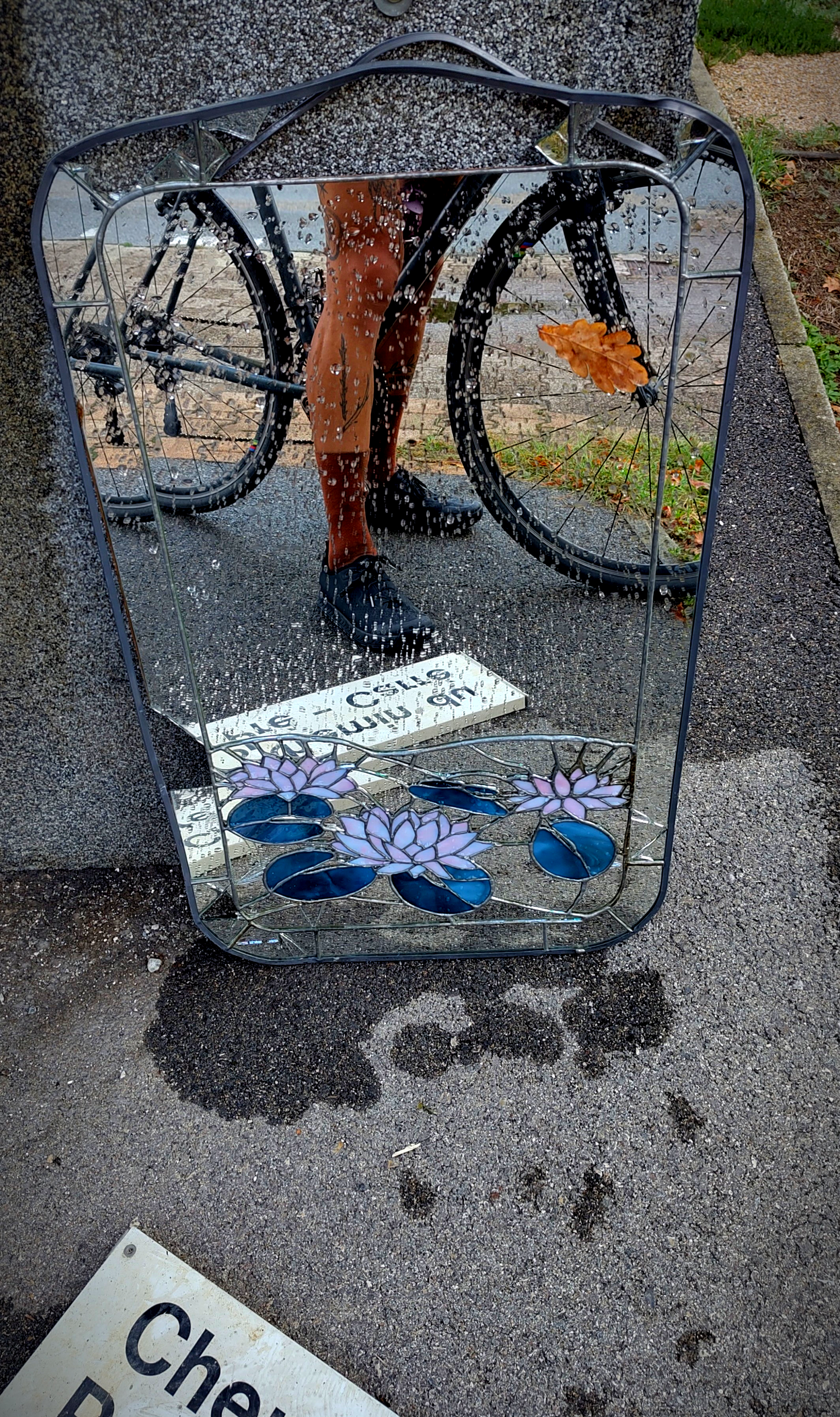 a picture of an art nouveau mirror partially broken abandonned against a wall reflecting a street sign that says 'chemin du pré...' and the photographer bicycle and legs