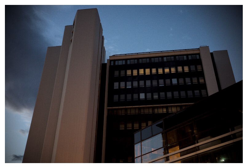 a tall building viewed from below in an early night light. the sky still retaining some of its blue hues. a grey shadow behind the building.