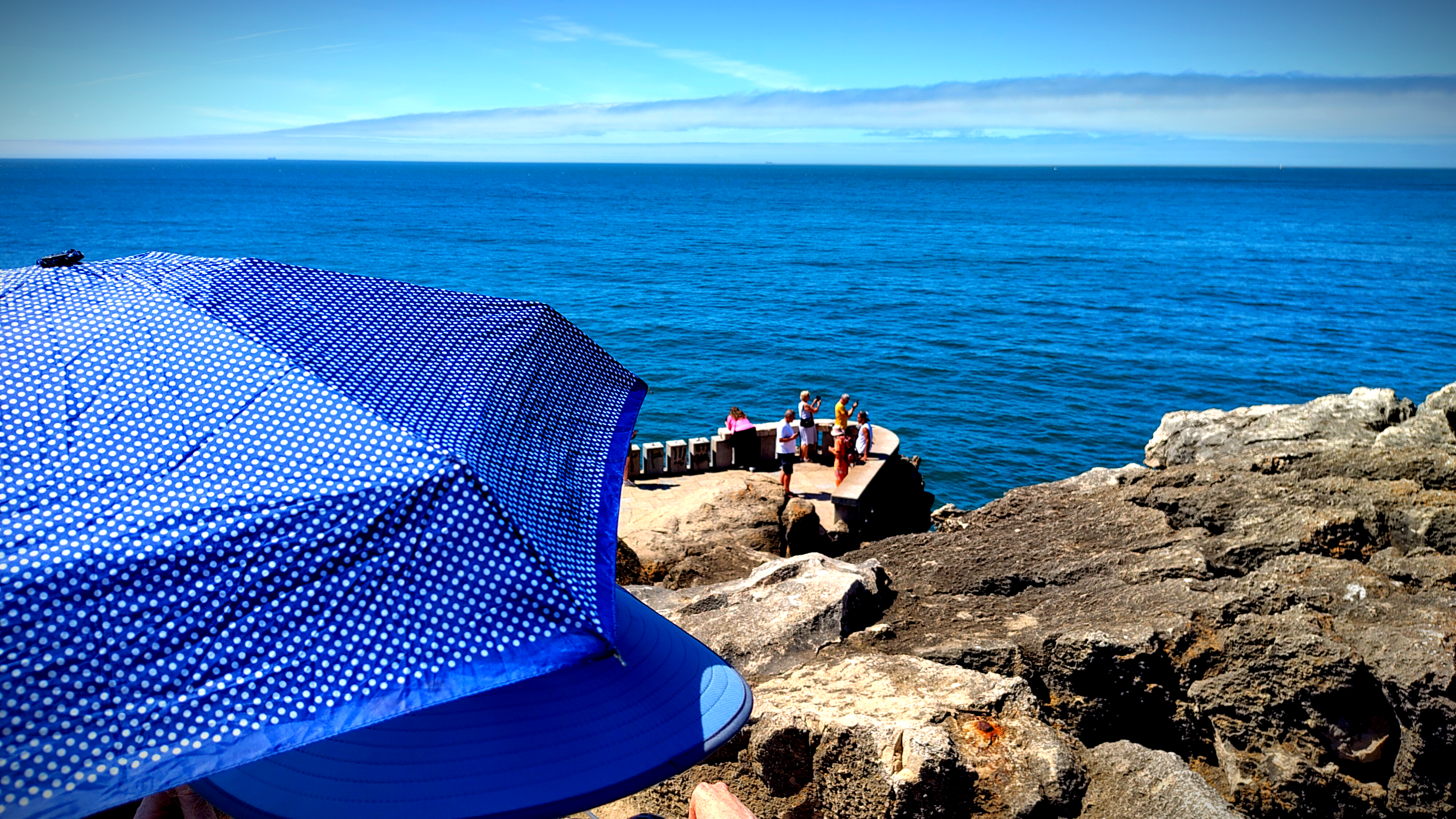 a picture of the ocean and the sky, on the bottom left corner a blue umbrella matches the color of the ocean and sky. on the right bottom corner rock cliffs and people photographing the view.