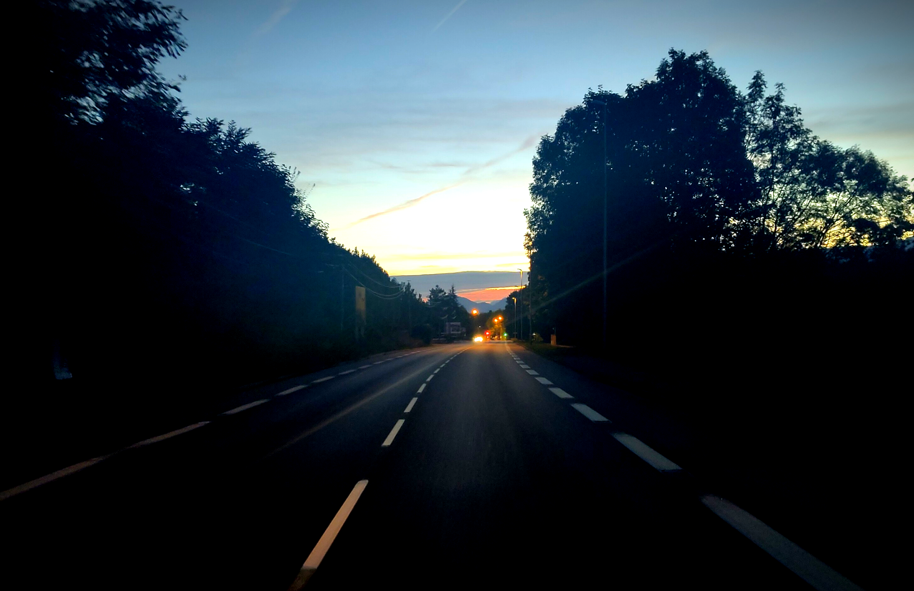 a barely lit asphalt road cornered by trees, it is a slight descent and on the horizon we see a car coming with its headlight and the sky turning red