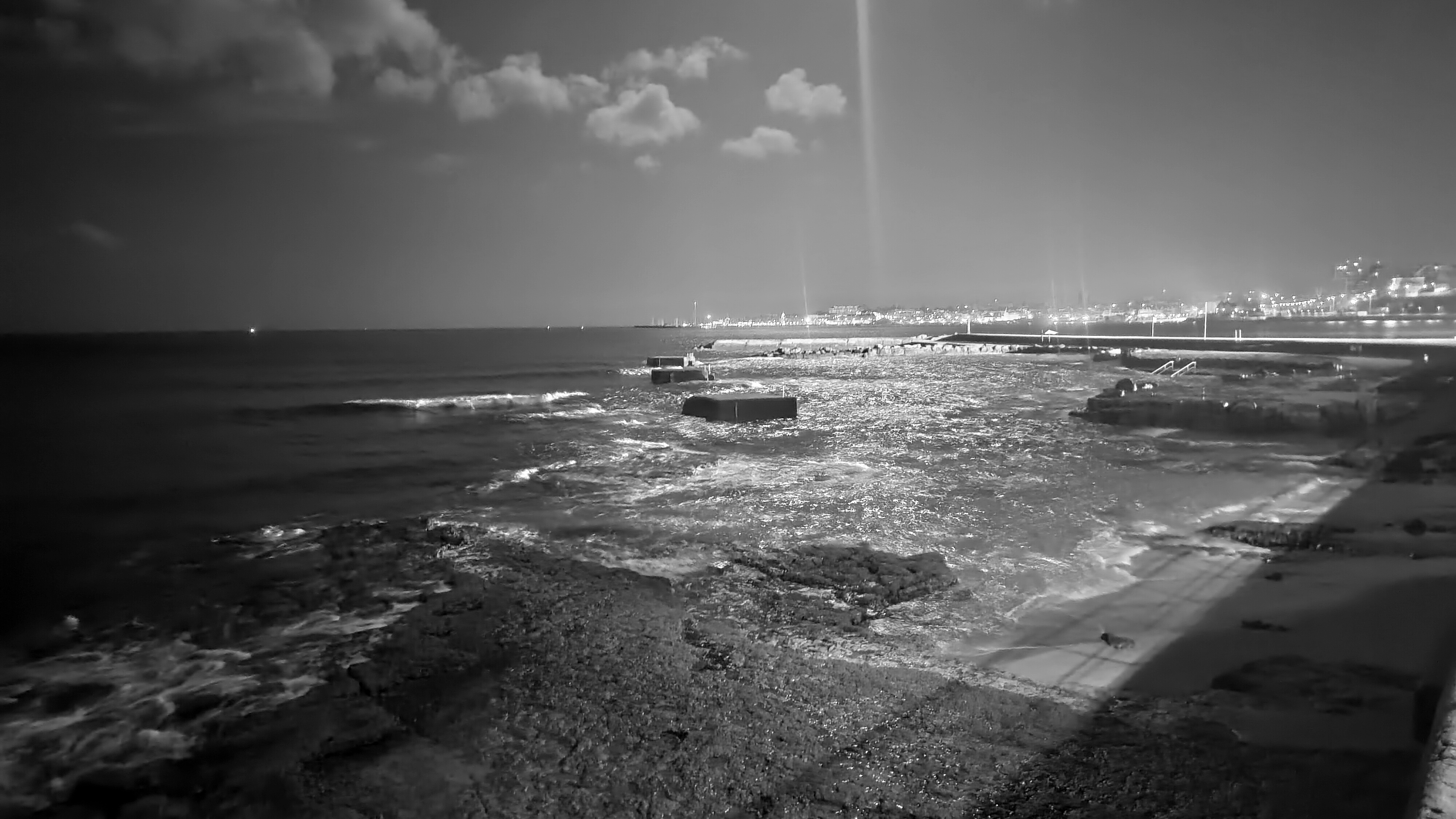 a tidal pool with waves shining with artificial light and moonlight. the sky is dark but you can see some clouds. far away lights of the city.
