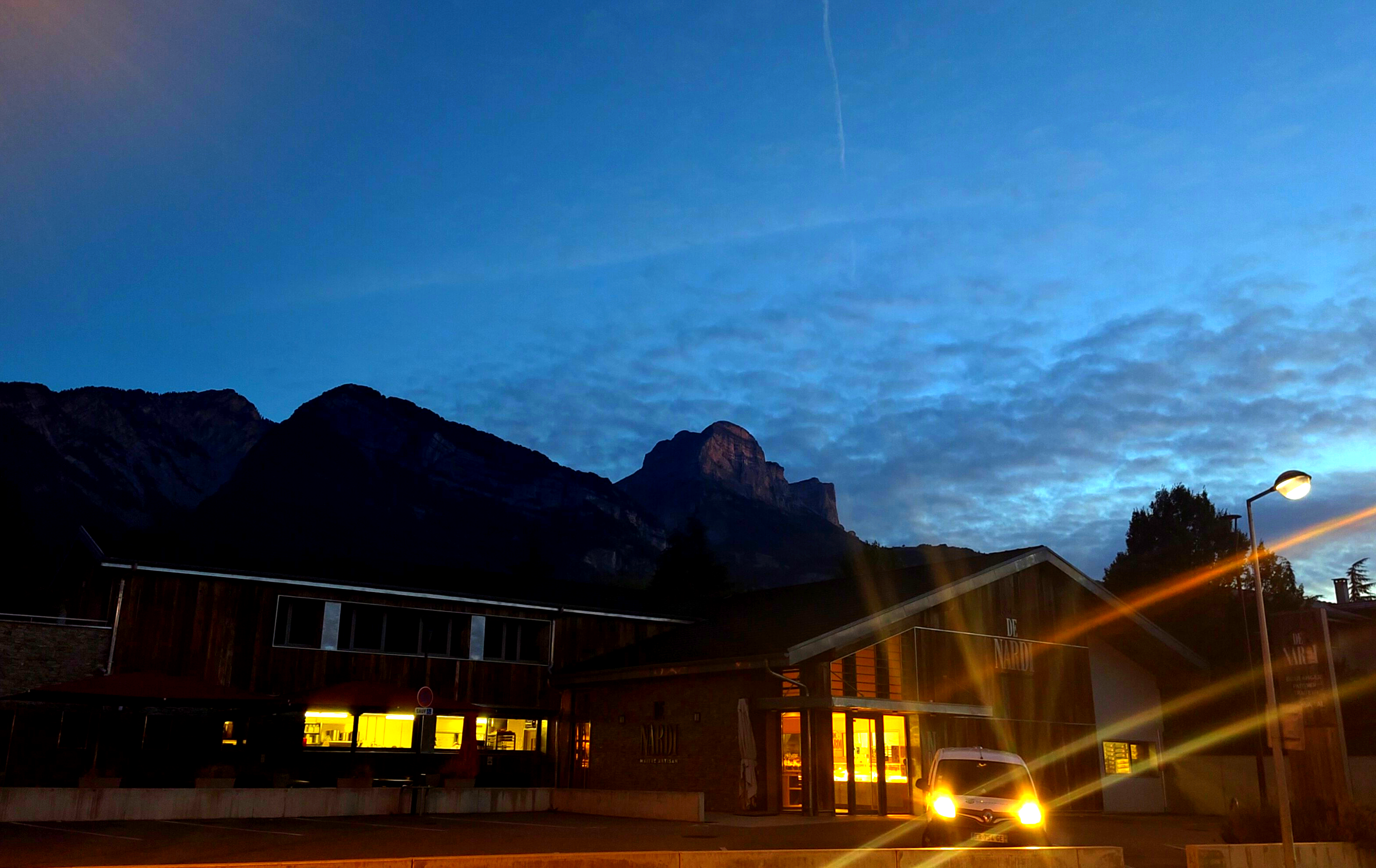 a night shot of the Dent Crolles mountain summit tinted red by the sunrise. below it, closer to the photographer a bakery with its lights on a parked car with headlights on